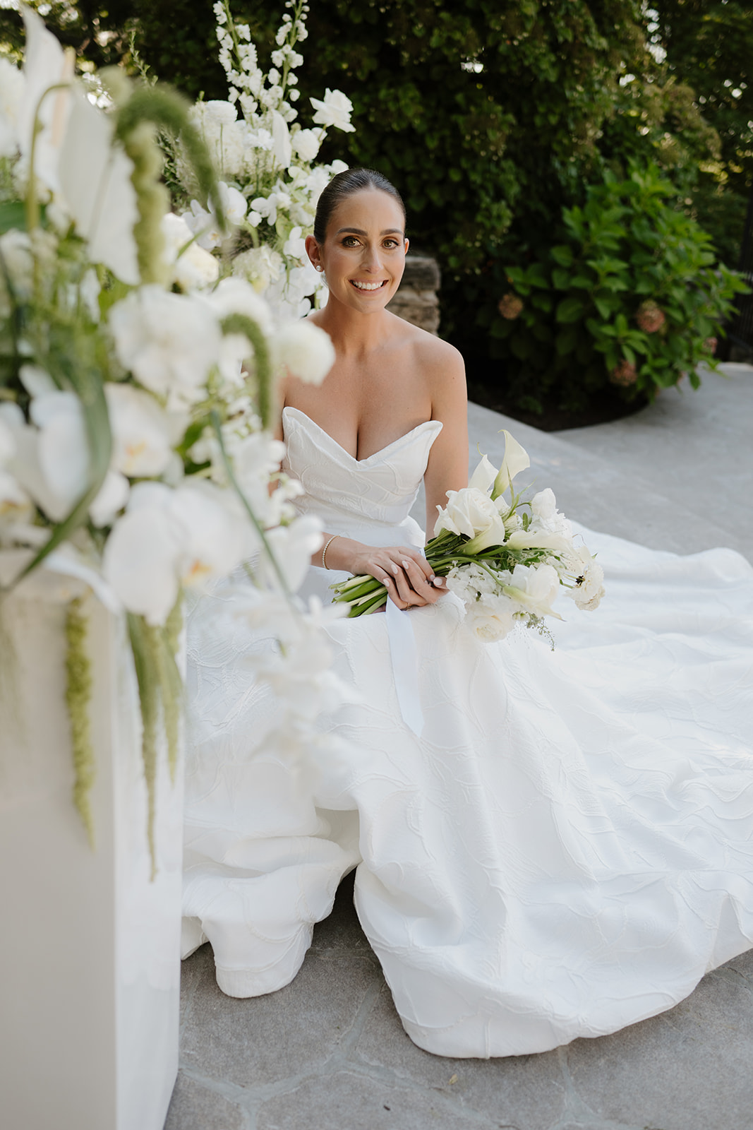 Bride sitting on stone steps with her gown spread out, smiling radiantly while holding her bouquet.